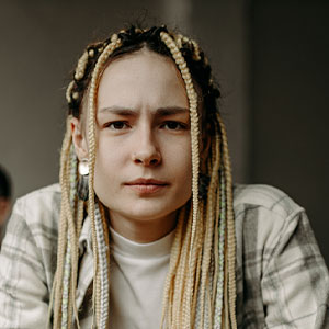 Jade is a Black or missed ethnicity woman with long bleached braids and black roots. She wears a white and green flannel over a white turtleneck.