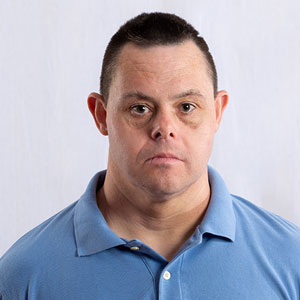 Jason is a White man wearing a blue polo shirt. He has black hair in a buzz cut. He has facial features of Down syndrome and is frowning.