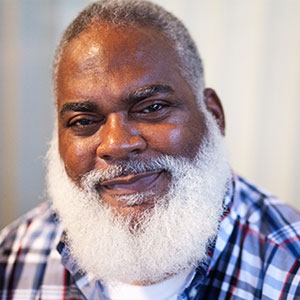 Marquis is a Black man with a closely shaved head and full white beard and mustache. He is smiling. He wears a red, white, and blue flannel shirt.